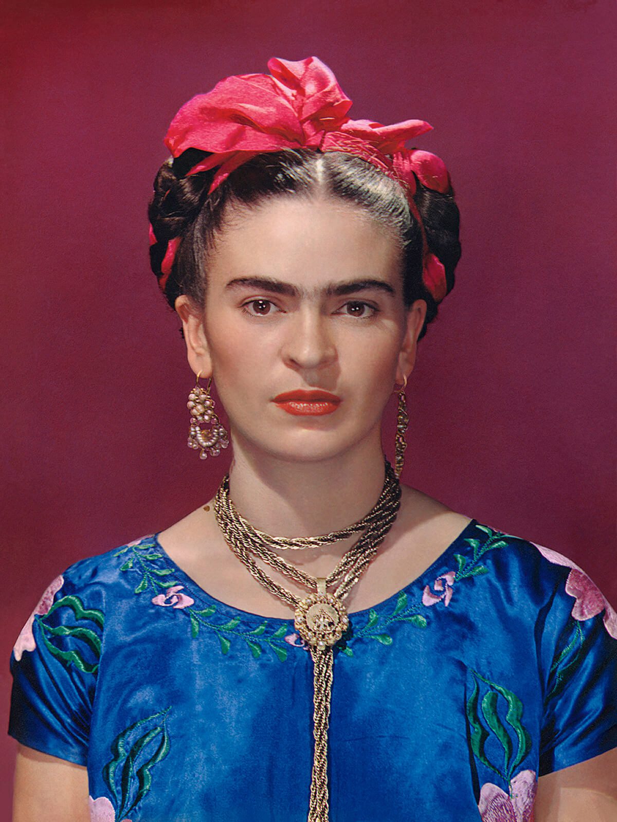 Mexican artist Frida Kahlo pictured in blue silk blouse standing against a maroon background