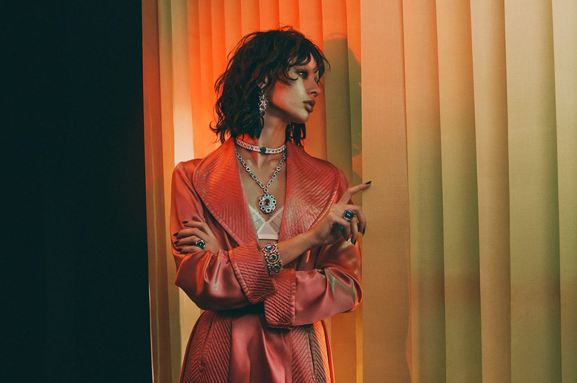 Model stands looking out of blinds wearing multiple jewels