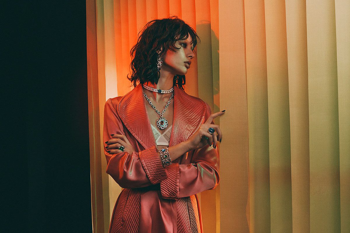 Model stands looking out of blinds wearing multiple jewels