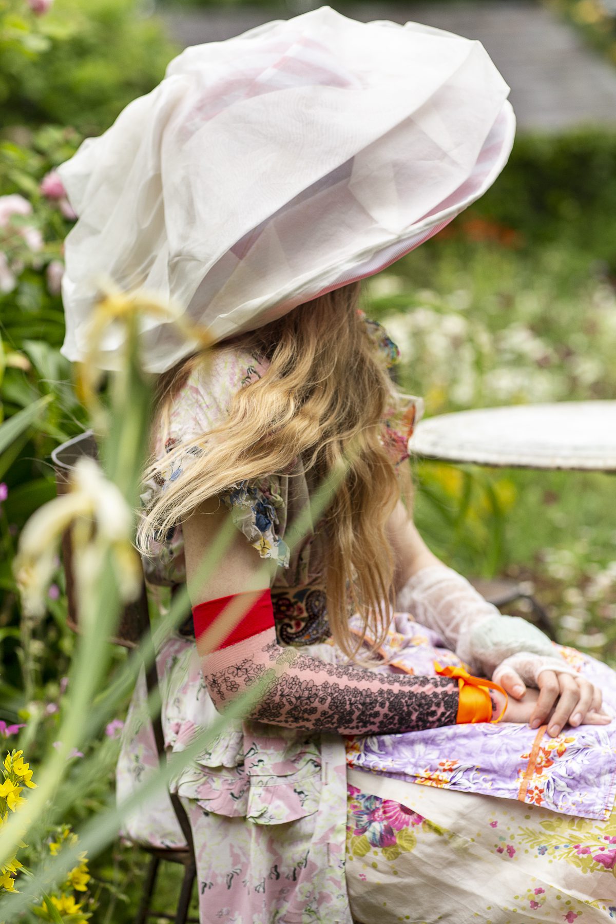 Model poses wearing a large hat seated in long grass field