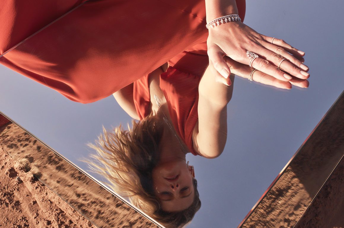 Model leaning over a mirror wearing a red dress and diamond jewellery
