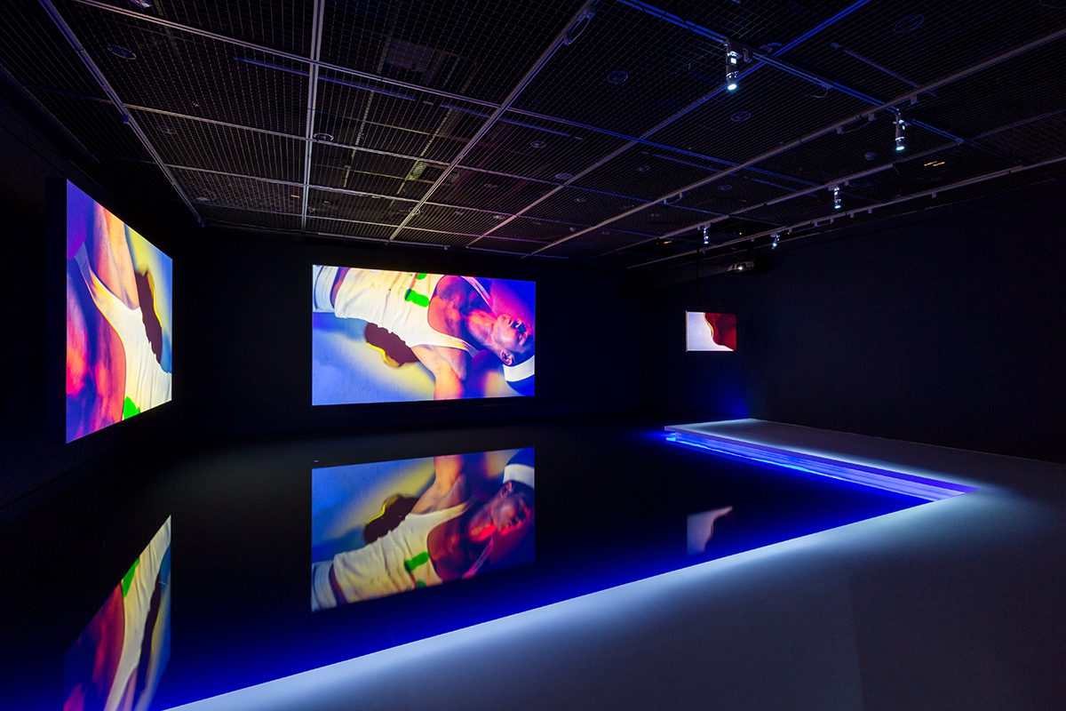 three screens with coloured lights in a dark room showing a man lying down wearing a vest