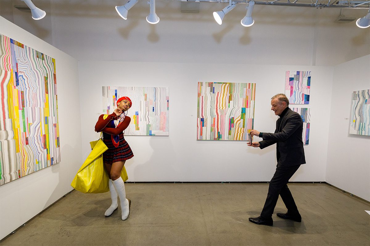 A girl posing in an art gallery and a man taking a photo on his IPhone