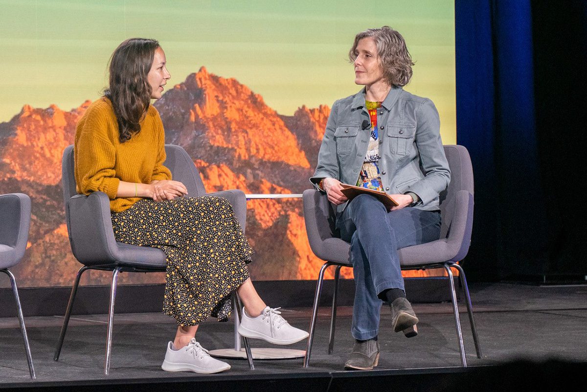 Two women speaking to each other sitting on chairs next to each other on a panel