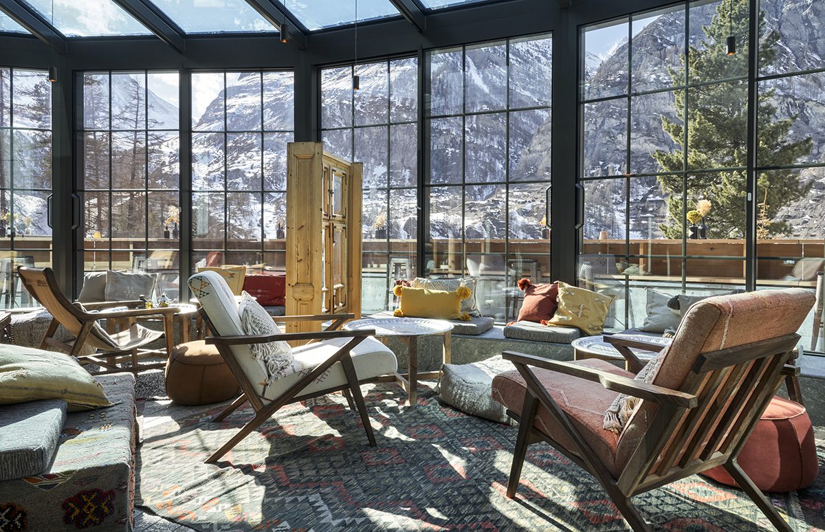 lounge chairs and deck chairs in a room with cushions and snow covered mountains outside the big windows