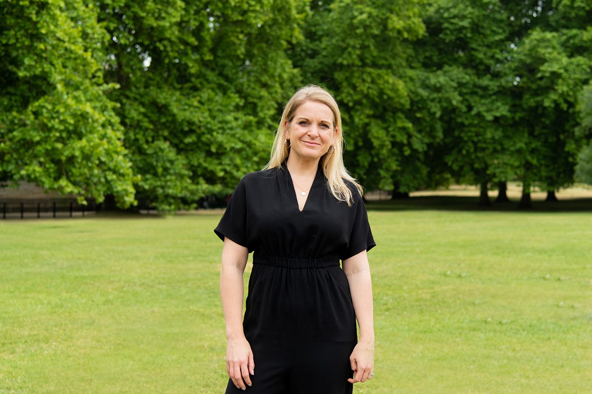 A blonde woman wearing a black dress standing in front on trees on the grass