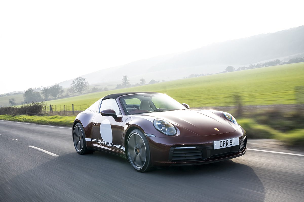 a red convertible Porsche with a white circle on the side of the car, driving by a green field