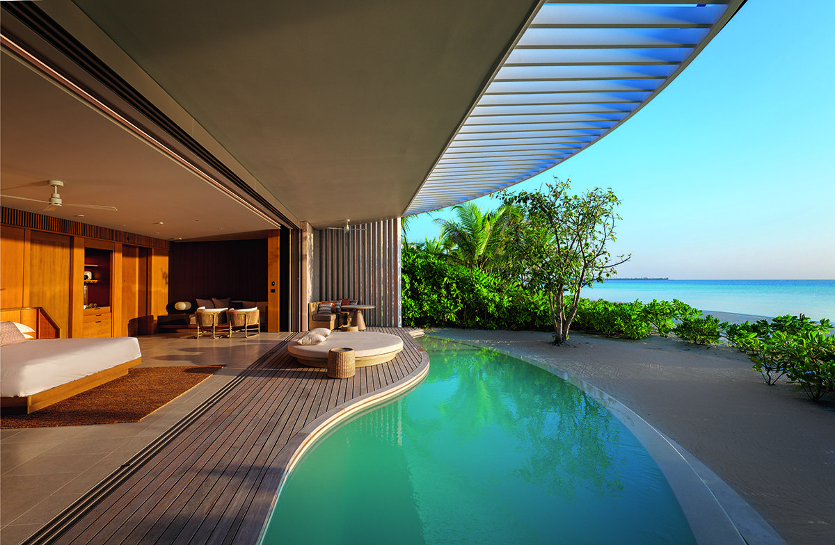 A bedroom leading to a swimming pool that leads to the sand on a beach with plants and trees