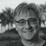 A man wearing glasses, with palm trees behind him