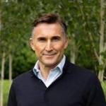 A man wearing a black v-neck jumper and shirt underneath standing on the grass with trees behind him