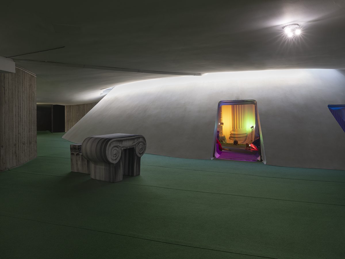 A room with a green carpet and top of a greek column as seat with an entrance to a dome inside it