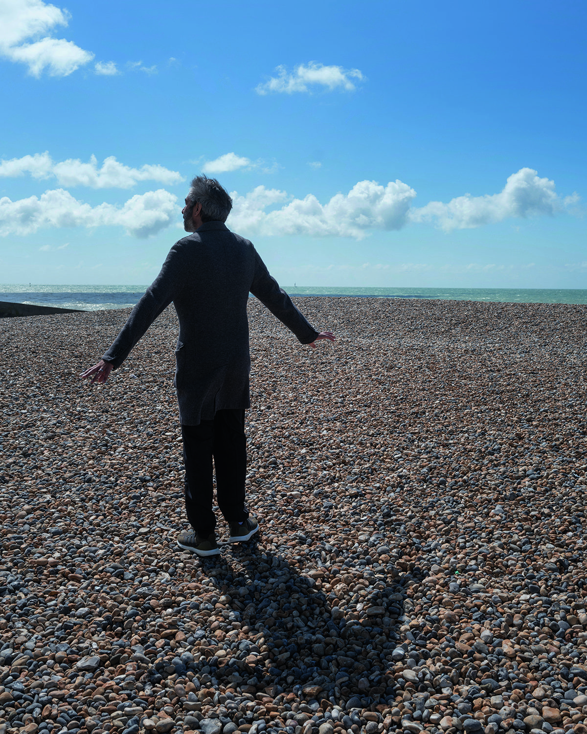 A man standing on a pebbled beach wearing a white t-shirt, black jeans and a long coat with his arms spread out