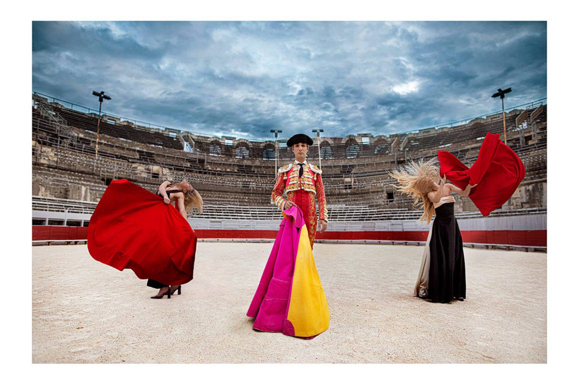 A man in a bullring holding a pink and yellow flag with women on either side of him holding red flags