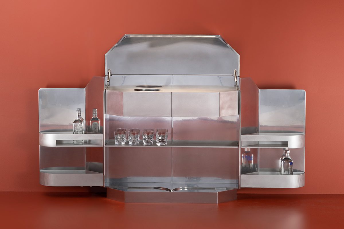 A silver metal bar with glasses in it in front of a red wall