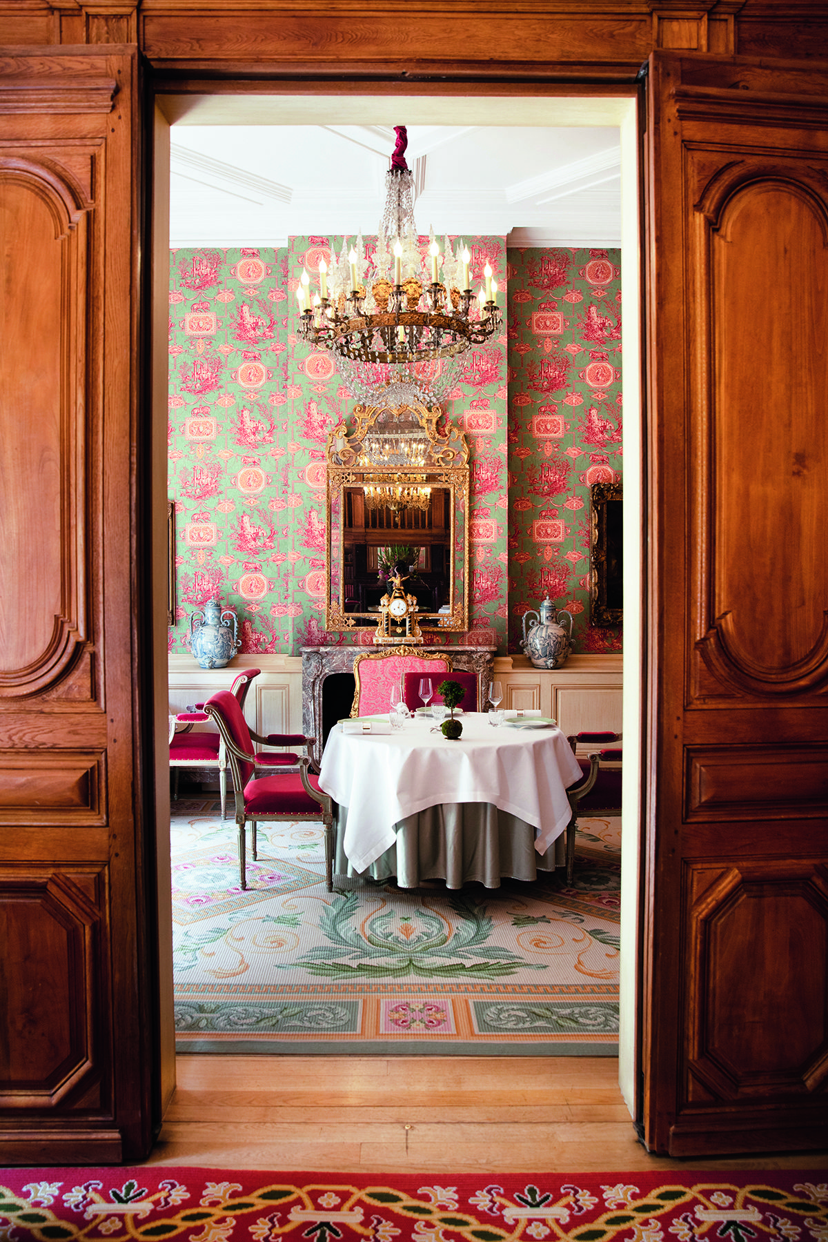 two wooden doors opening to a room with a red chair and table with chandelier hanging over it