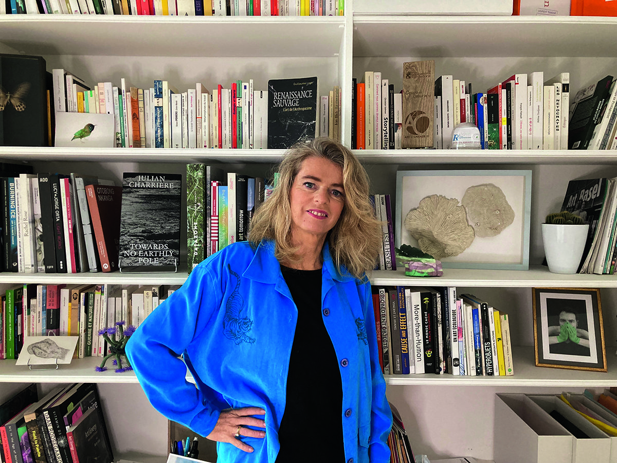A blonde woman in a black top and blue shirt standing by a book shelf with her hand on her hip