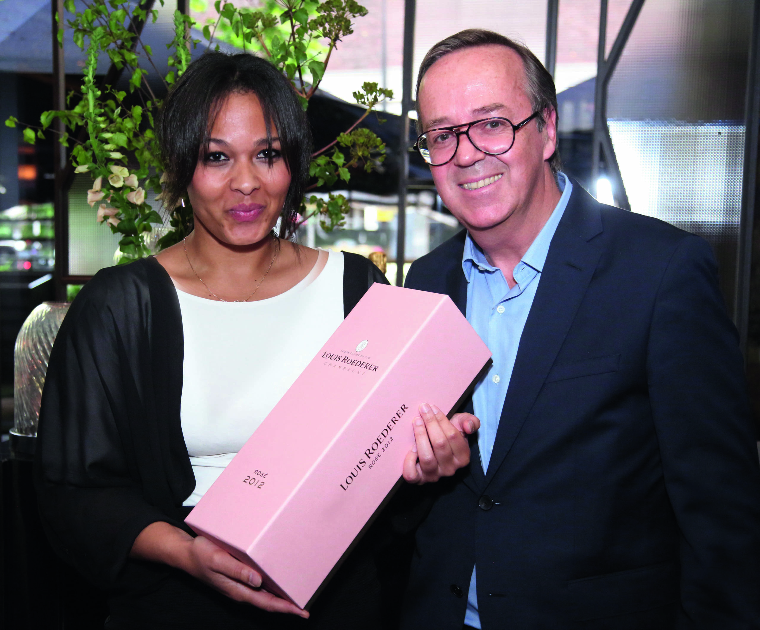 Woman in a white and black top holding a pink case of champagne standing next to a man in a blue shirt and black blazer