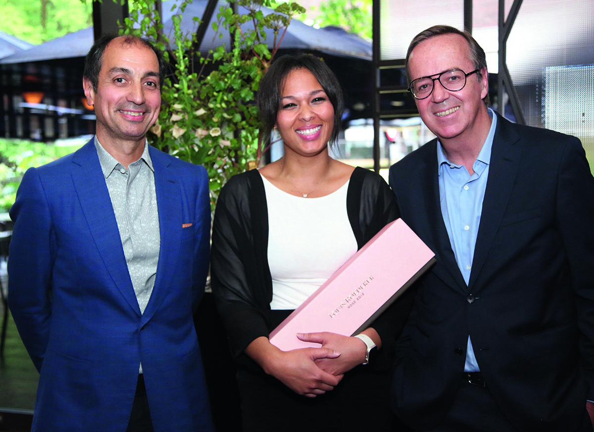 Woman in a white and black top holding a pink case of champagne standing next to two men in shirts and blazers
