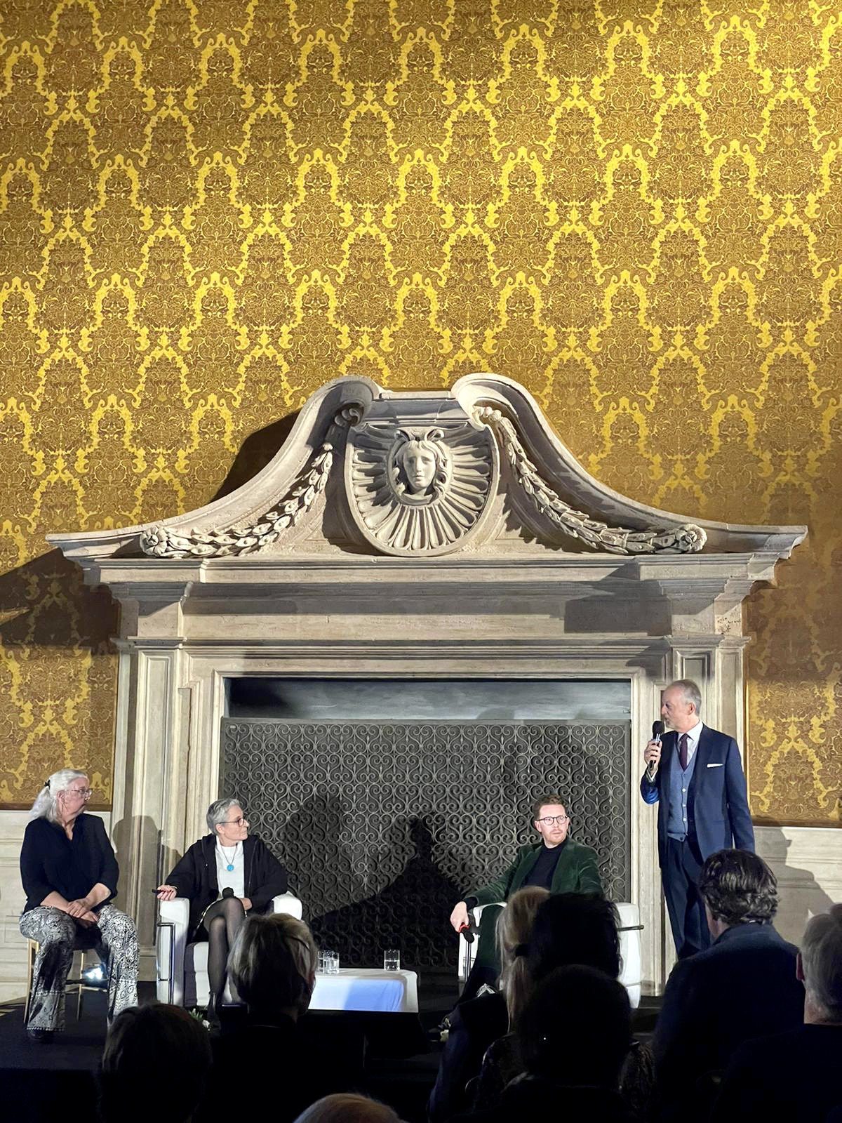 5 people sitting in front of marble structure and yellow patterned walls giving a talk and a host standing beside them with a microphone