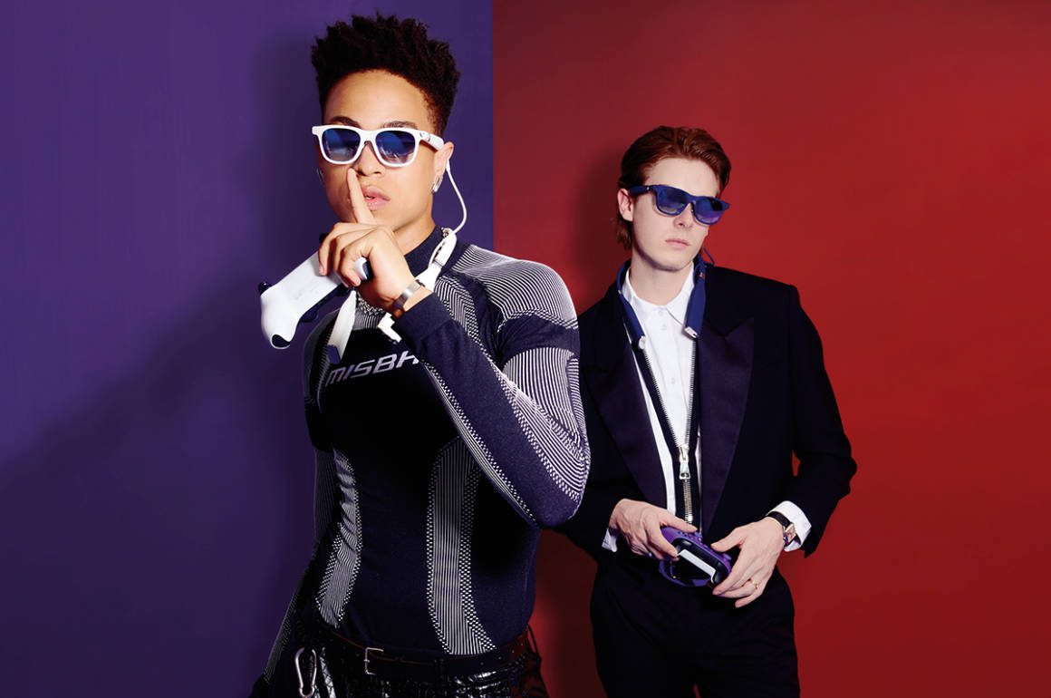 purple and red background with a model with his finger to his lips in a leather outfit wearing glasses and a man wearing a suit in the background also wearing glasses