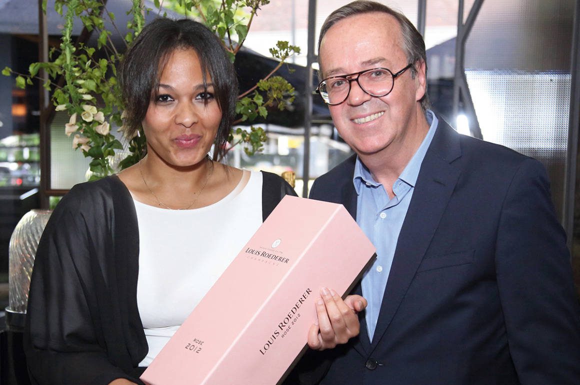 Woman in a white and black top holding a pink case of champagne standing next to a man in a blue shirt and black blazer