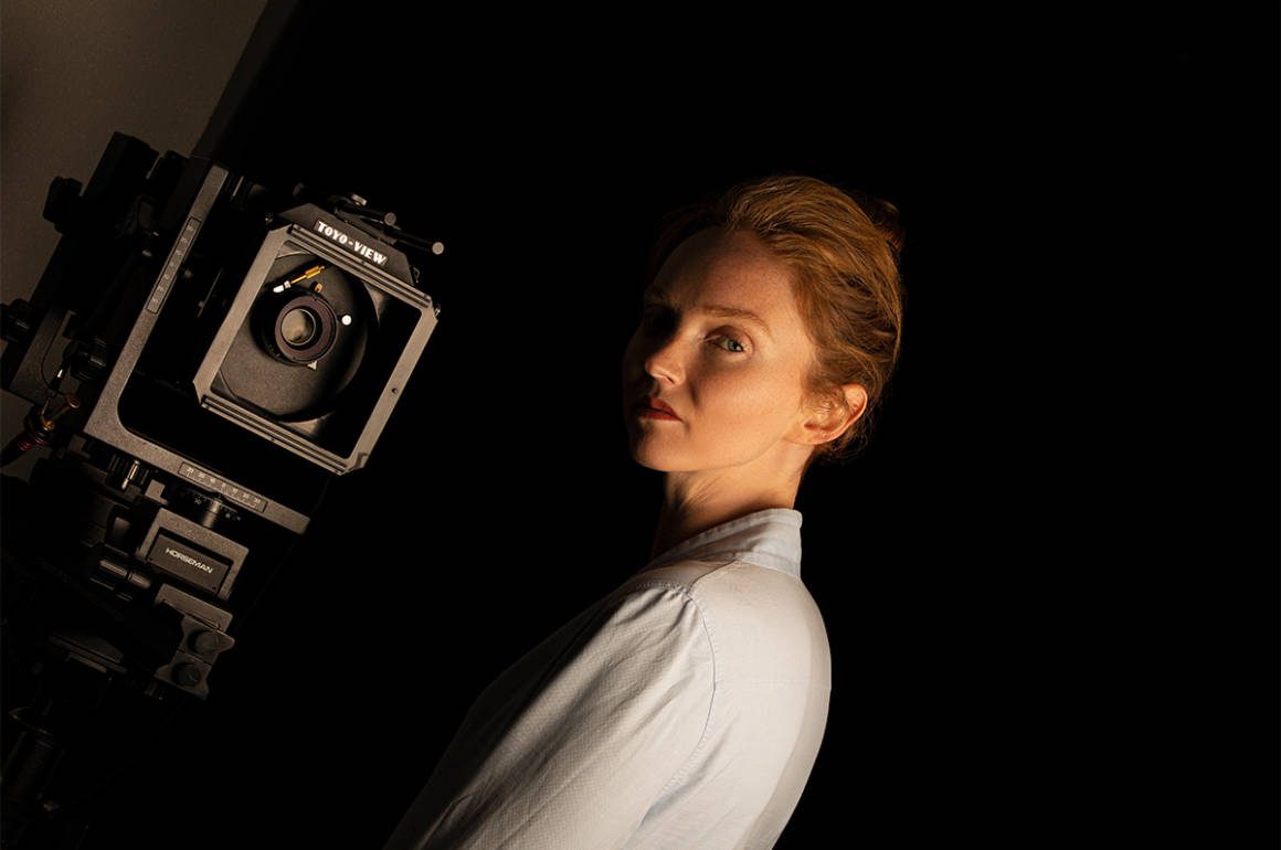 A ginger model with a wearing a white shirt with a camera next to her head