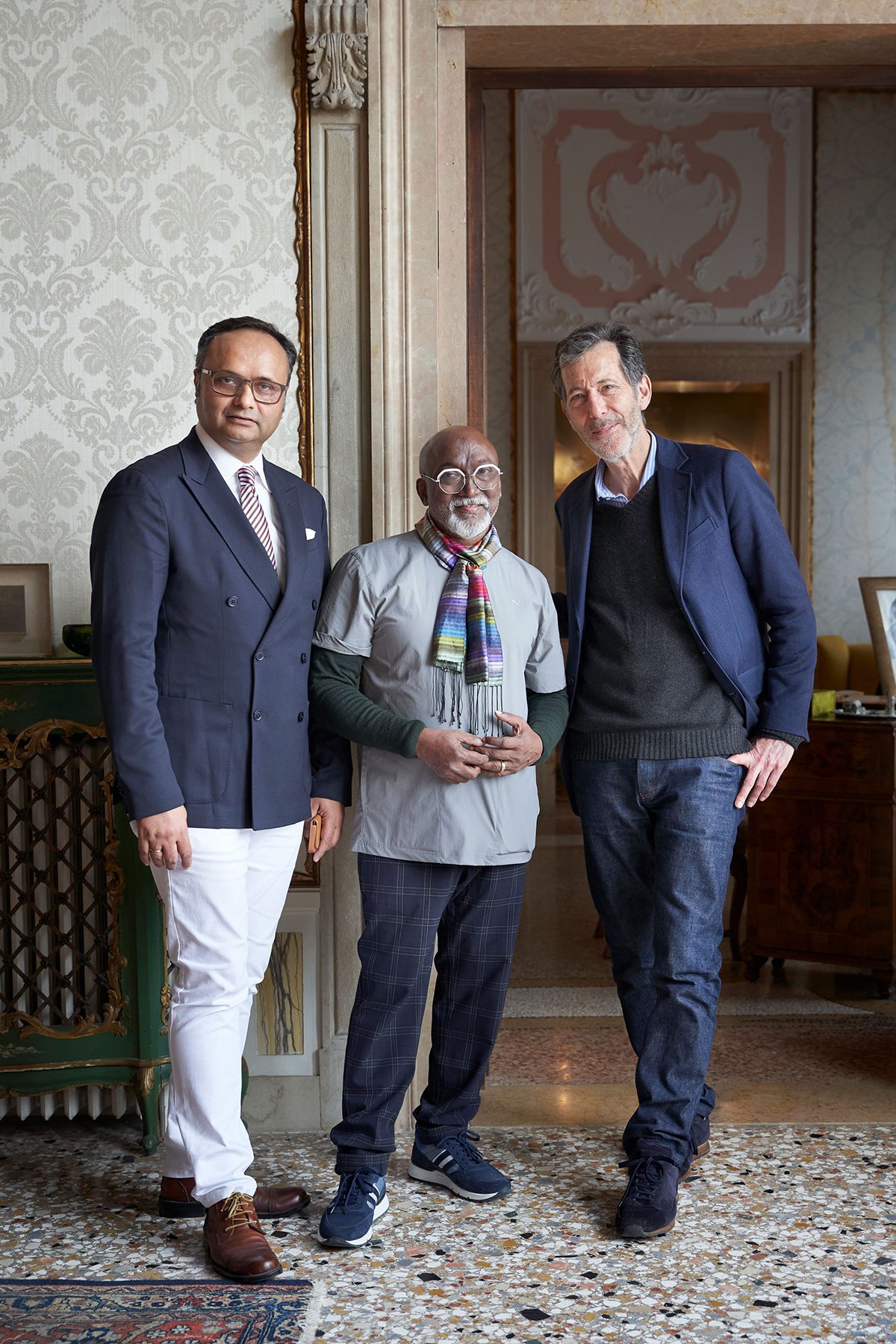 three men standing together, two in navy blazers and a smaller man in the middle wearing a multicoloured scarf and grey t-shirt