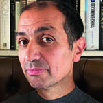 A man in a grey top sitting on a brown chair with books behind him