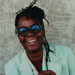 A woman laughing wearing blue sunglasses and a green jacket with a patterned blue and pink scarf