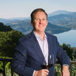 A man standing with a wine glass on a balcony with a lake and vineyards in the distance