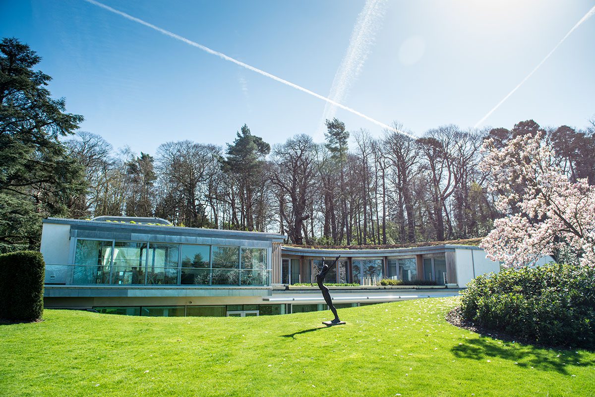 A blue glass building on the grass surrounded by trees and a statue in front of it