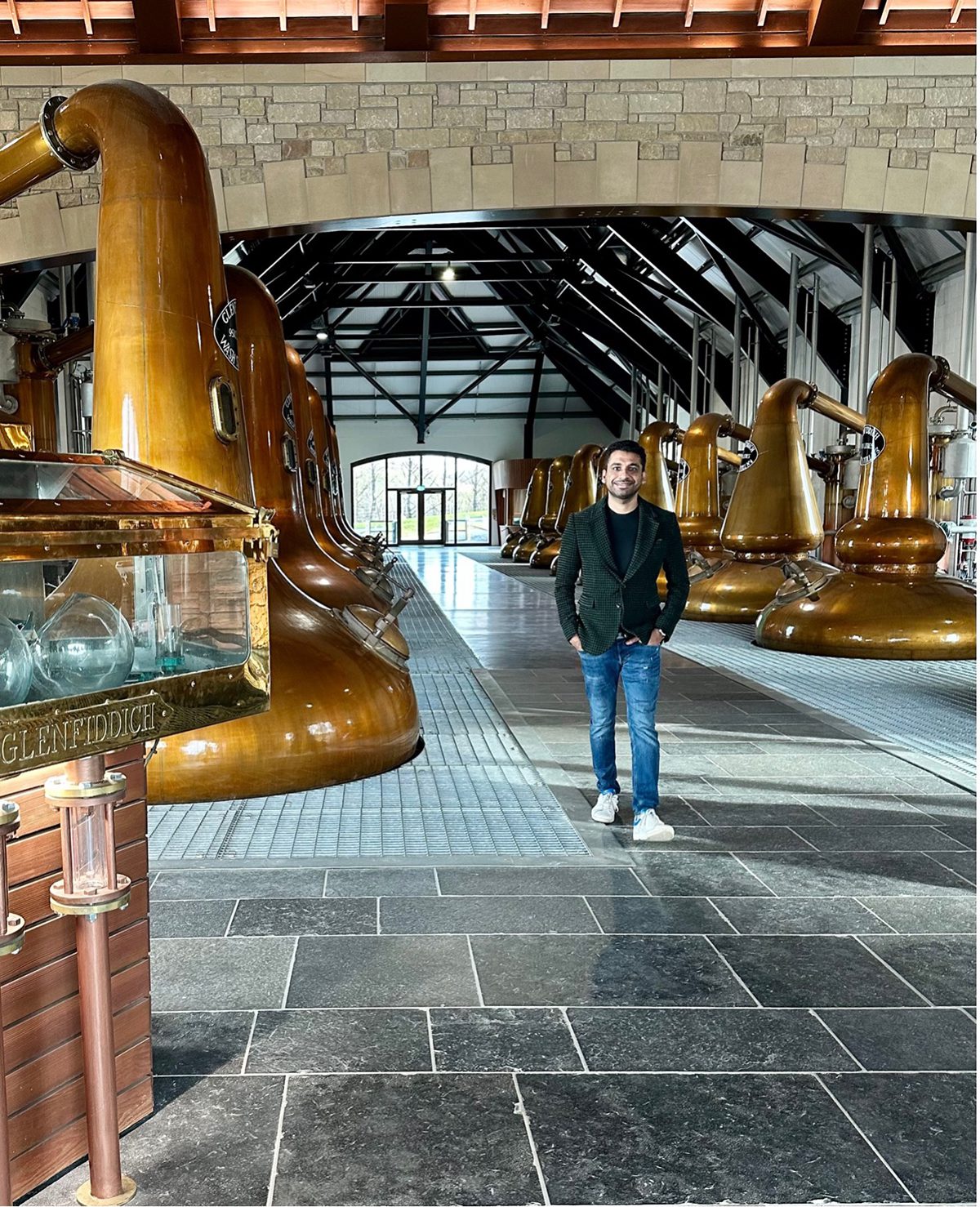 A man standing in a whiskey factory with large gold bells wearing jeand and a grey jacket
