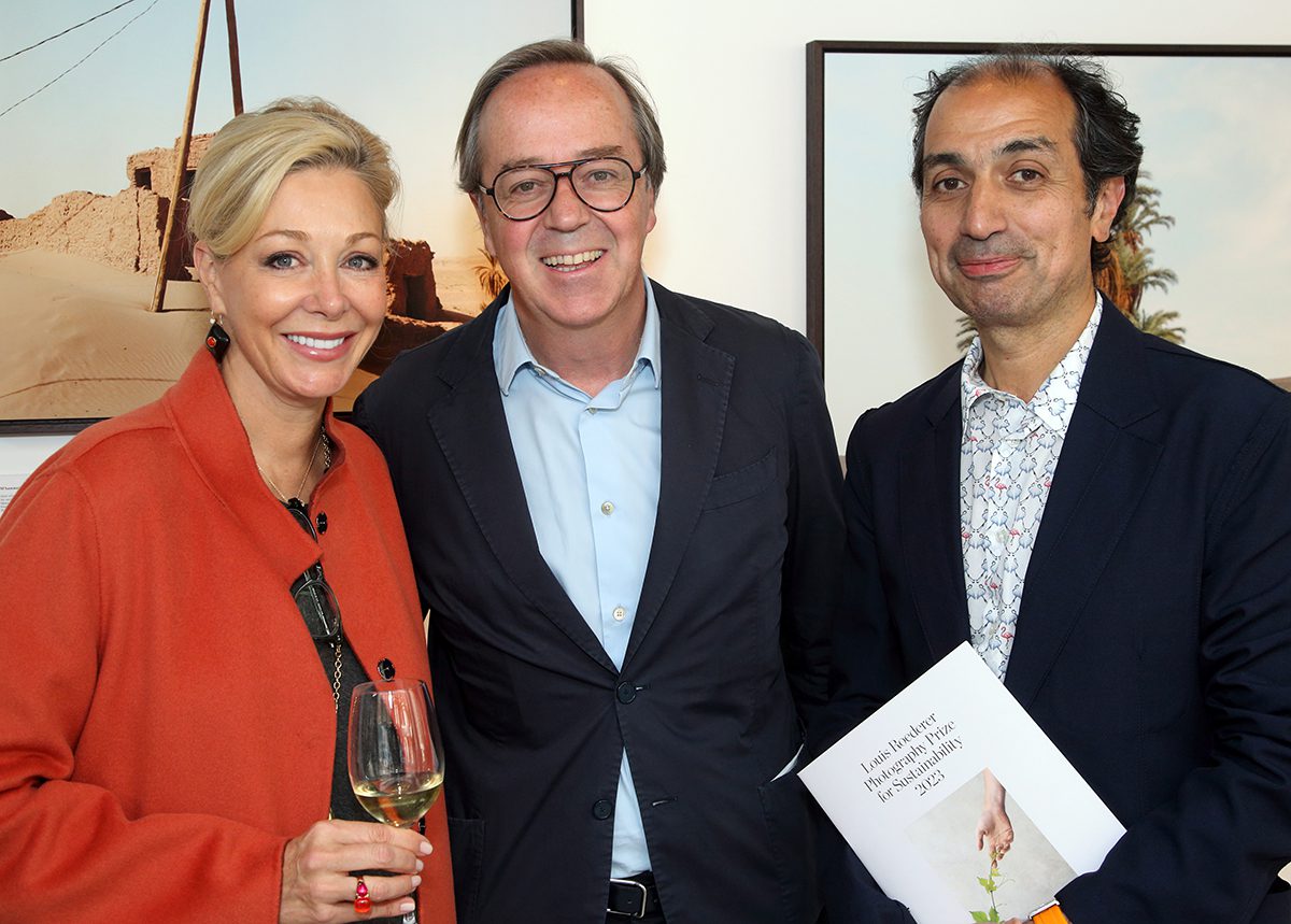 A woman wearing a red coat holding a glass of champagne standing next to two men in shirts and blazers