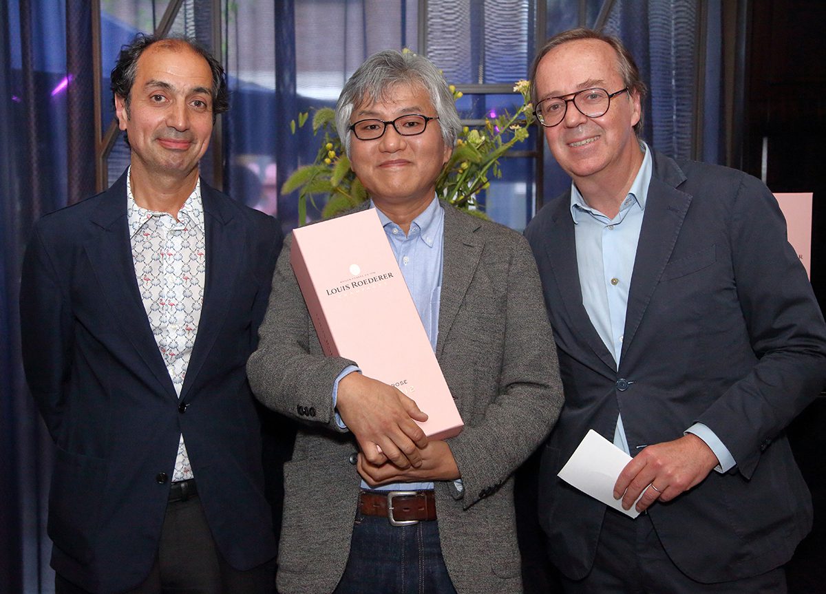 Three men wearing suits and the man in the middle holding a pink case of champagne