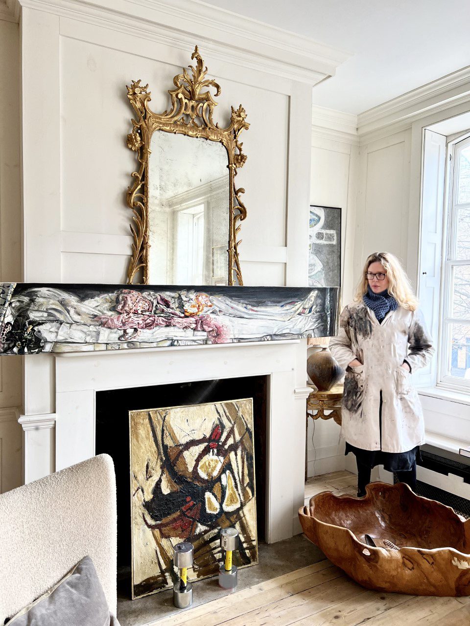 A woman standing by a chimney in a dirty white jacket with art works around her
