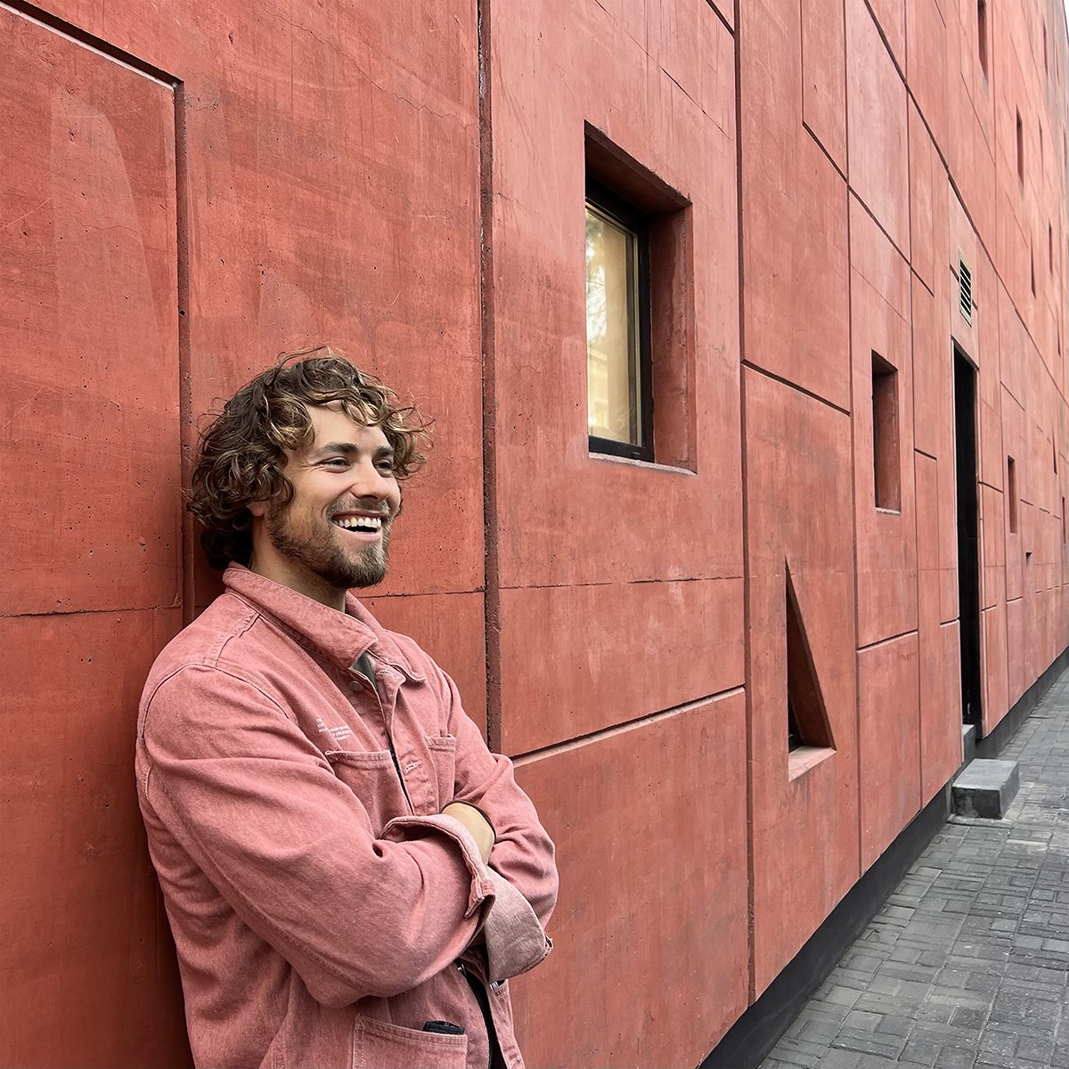 a man wearing a pink jacket standing by a pink wall with his arms folded