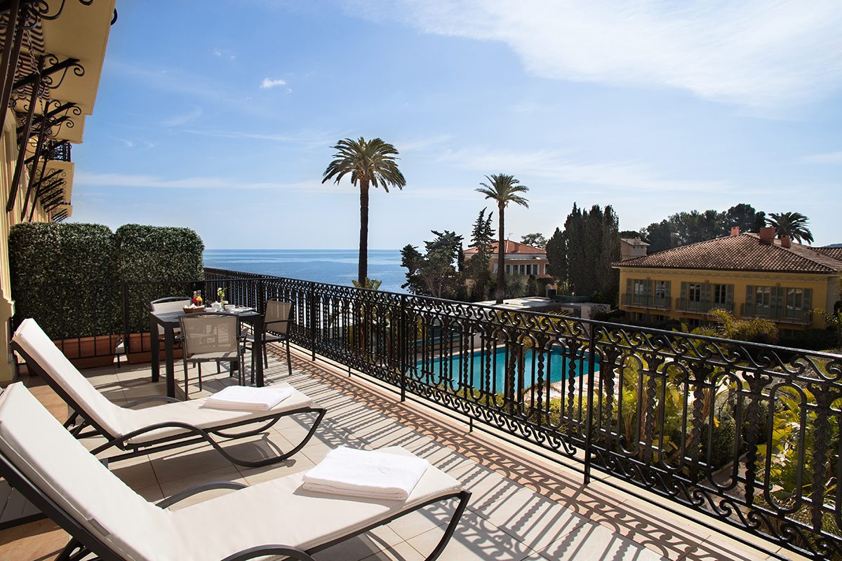 A terrace with deckchairs looking over a pool with palm trees and the sea