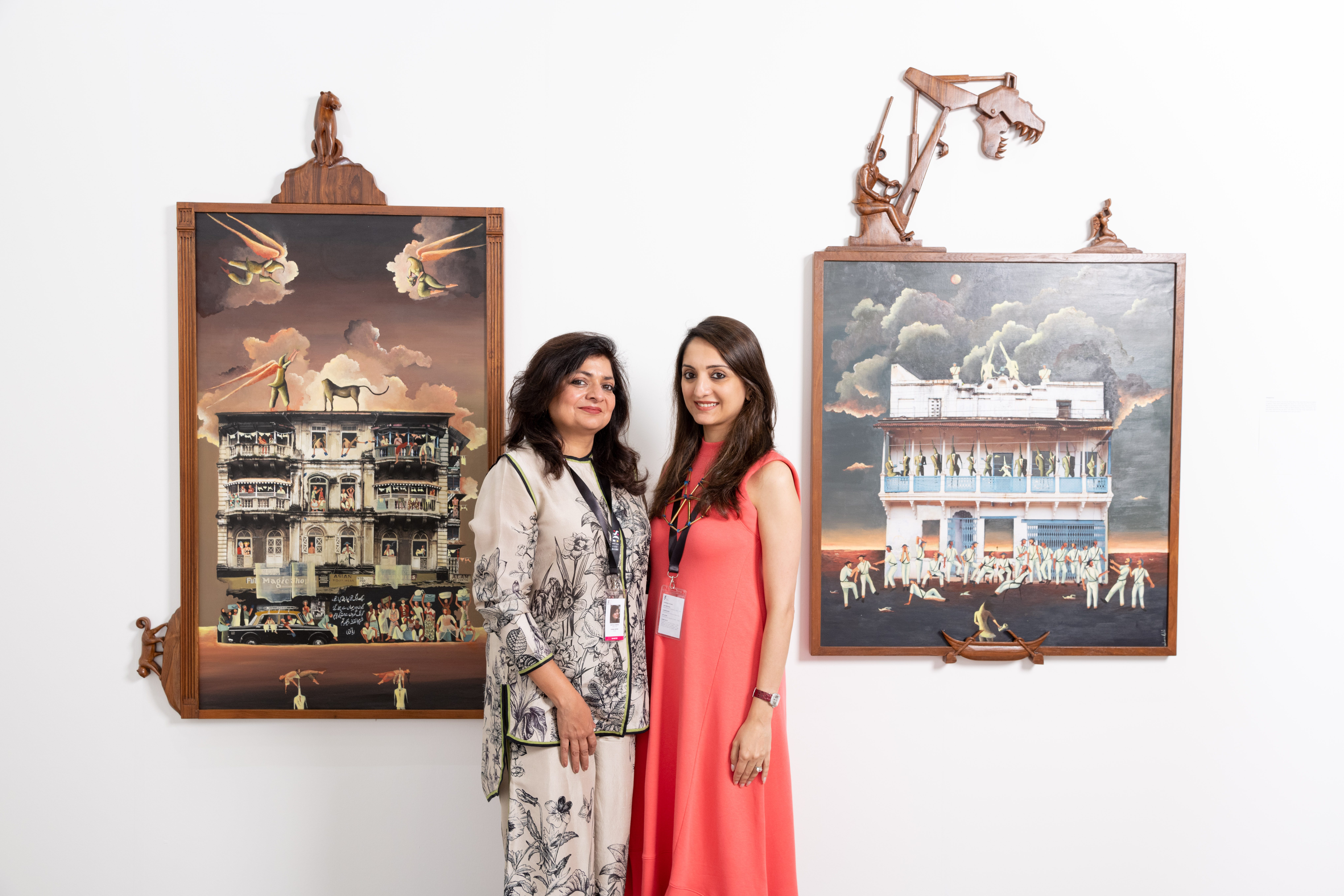 A woman wearing an orange dress standing next to a woman in a grey and white outfit standing between two paintings on walls