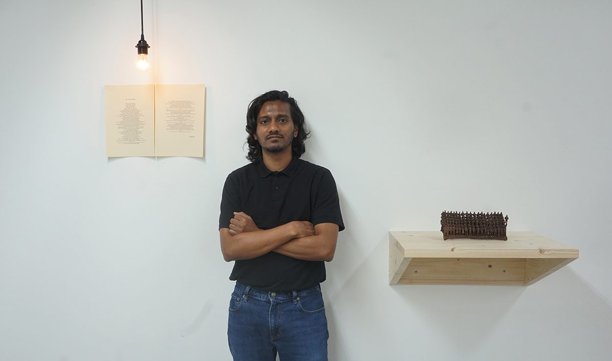 Man in a black t shirt and blue jeans standing with arms crossed by a white wall