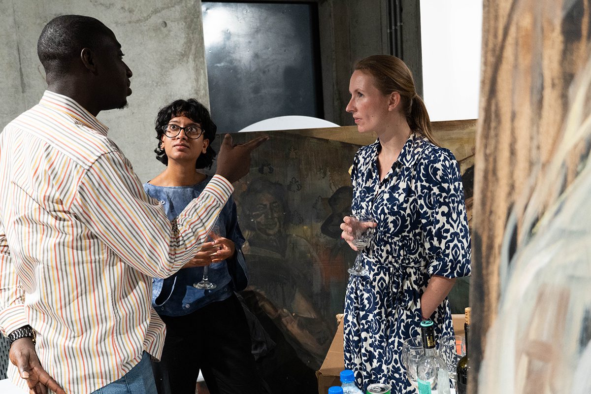 Three people speaking including a woman wearing a black and white dress and a man wearing a striped yellow shirt