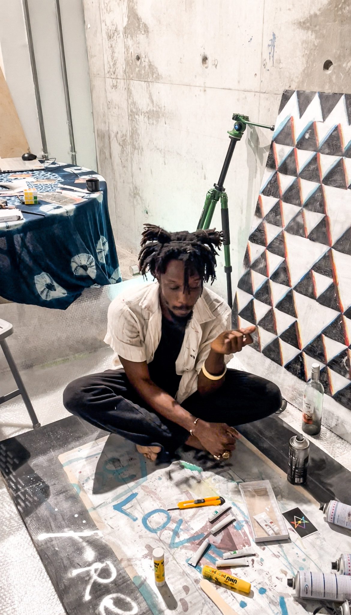 A man wearing black trousers and a white short sleeve shirt with a black top underneath sitting on the floor with a geometric picture beside him