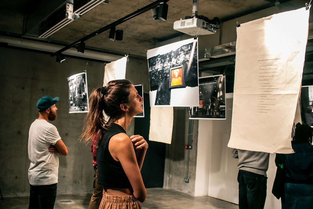 A woman with a pony tail looking at a work of art hanging on a string with a man beside her looking at another work on the string wearing a green cap