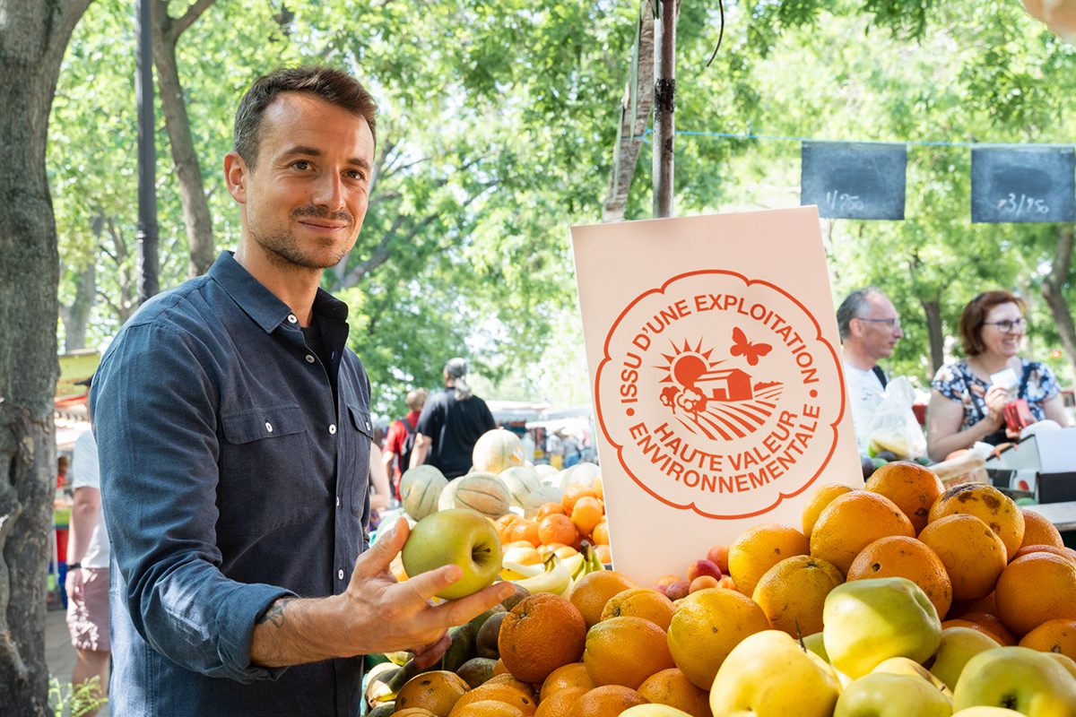 A man wearing a blue shirt holding fruit in his hand standing by a crate of fruit