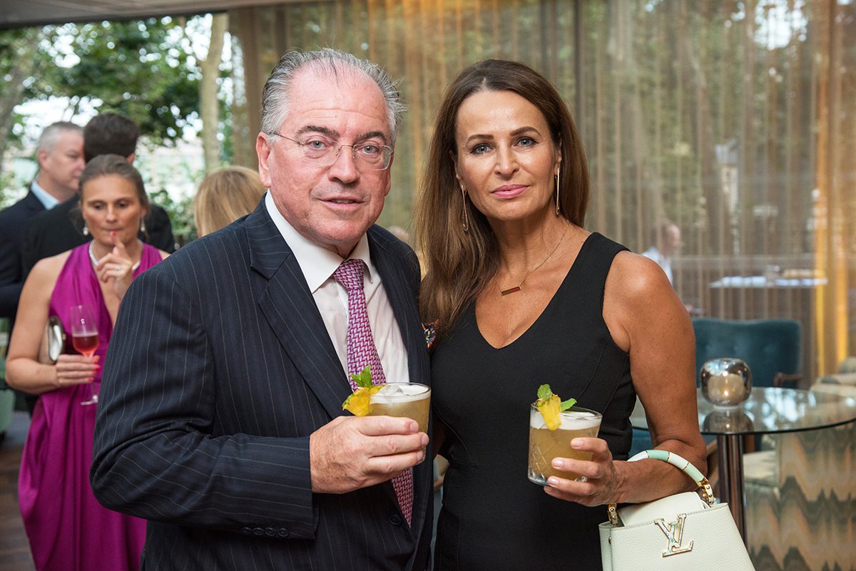 A man in a suit holding a drink next to a woman wearing black dress also holding a drink