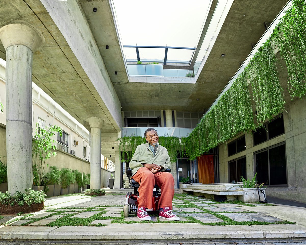 A man sitting in a wheelchair, wearing red trousers, in a courtyard of a building with plants hanging over the interior balconies