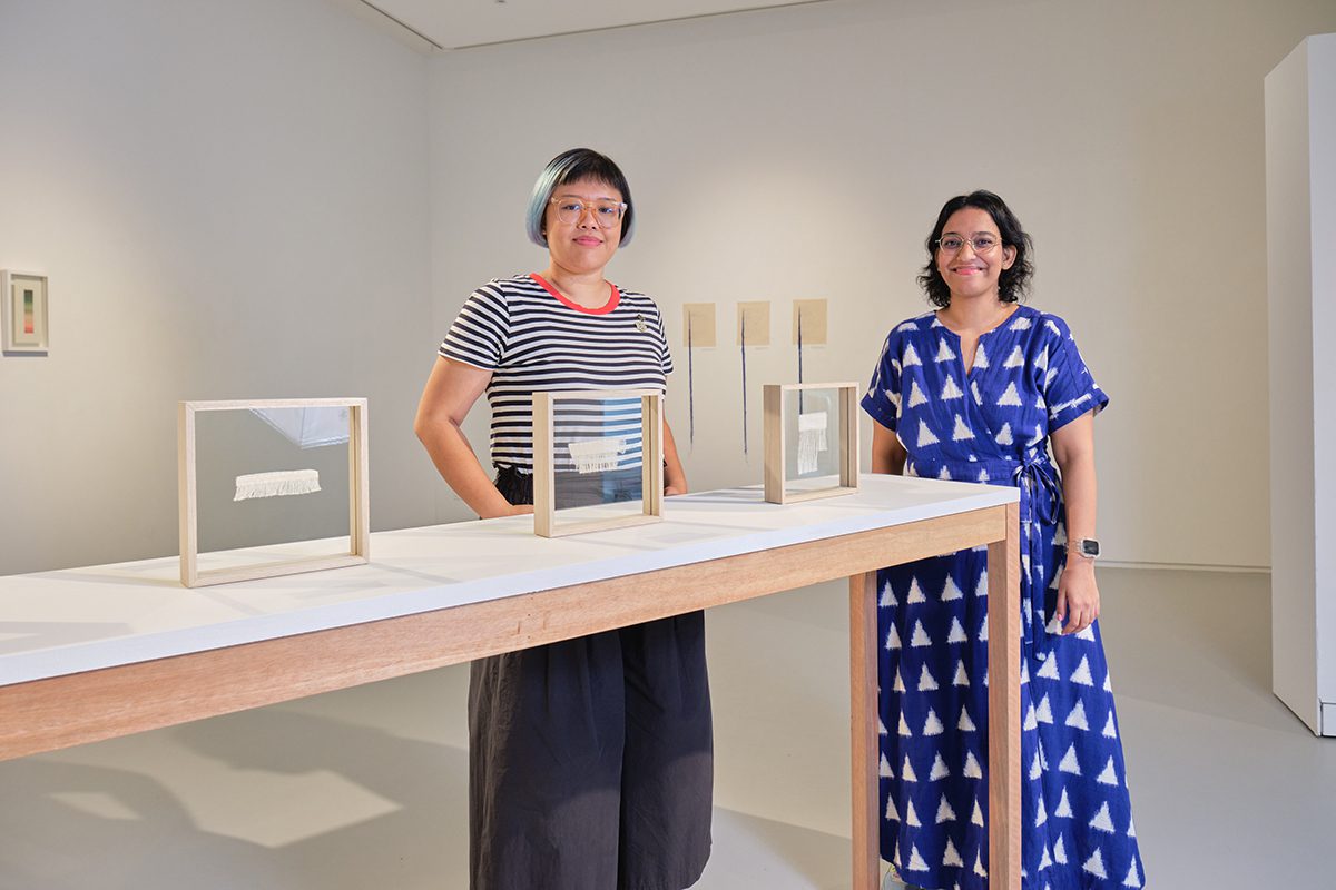 Two women standing by a wooden table with objects in glass frames on the table