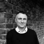 A man wearing a white shirt and black jumper standing by a brick wall