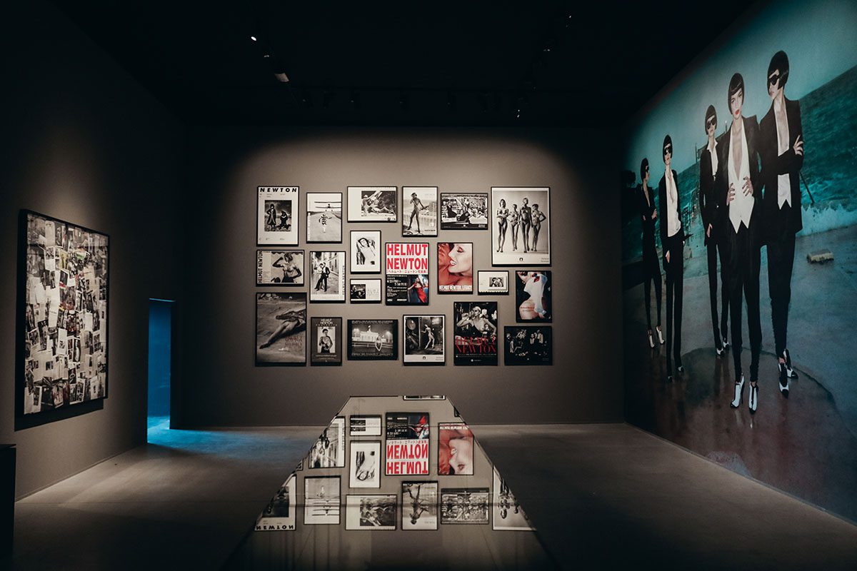 photographs hanging on a wall with Helmut Newton in white a ona. red background witten on a wall and a projected image on an adjacent wall
