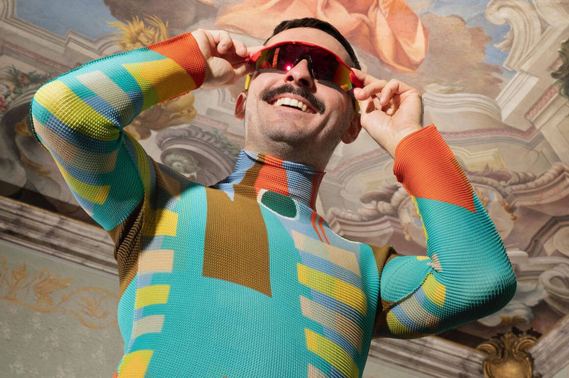 A man wearing a green, yellow and purple colourful top putting on red sunglasses