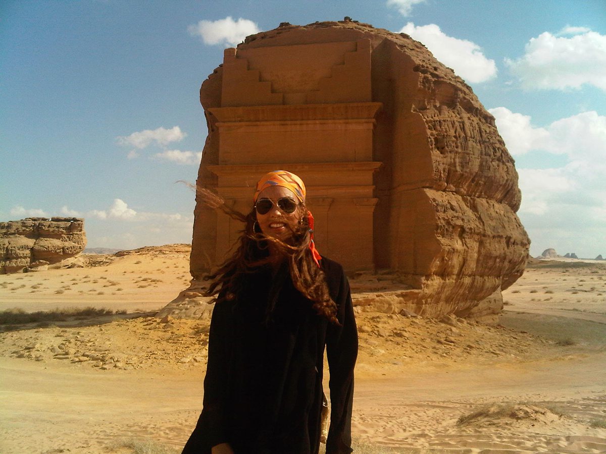  A woman wearing a black dress and orange head scarf standing next to a large rock in a desert